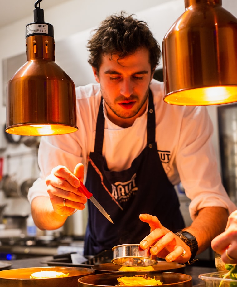 A chef plating a dish at the pass under warm heat lamps